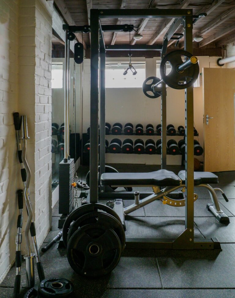 Compact home gym setup featuring a power rack with a bench, barbell, weight plates, cable pulley system, dumbbell rack, and multiple barbells against a white brick wall.