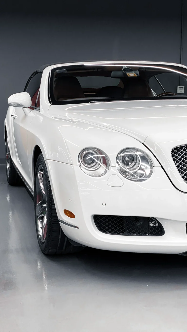A close-up view of the front end of a white Bentley Continental GT, showcasing its glossy paint, mesh grille, dual circular headlights, and chrome accents inside a showroom.