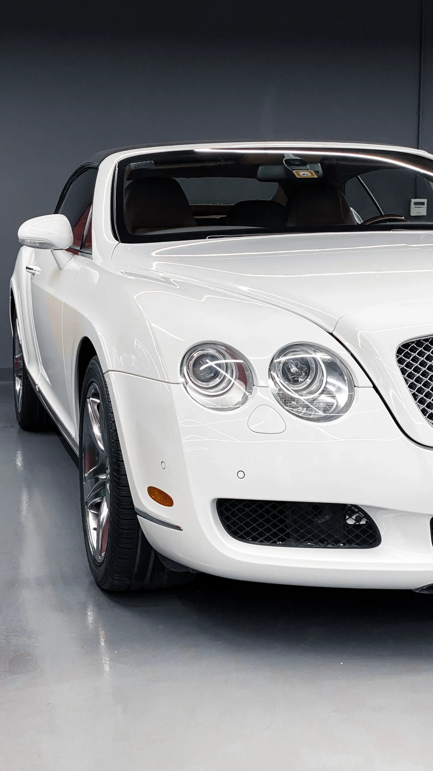 A close-up view of the front end of a white Bentley Continental GT, showcasing its glossy paint, mesh grille, dual circular headlights, and chrome accents inside a showroom.