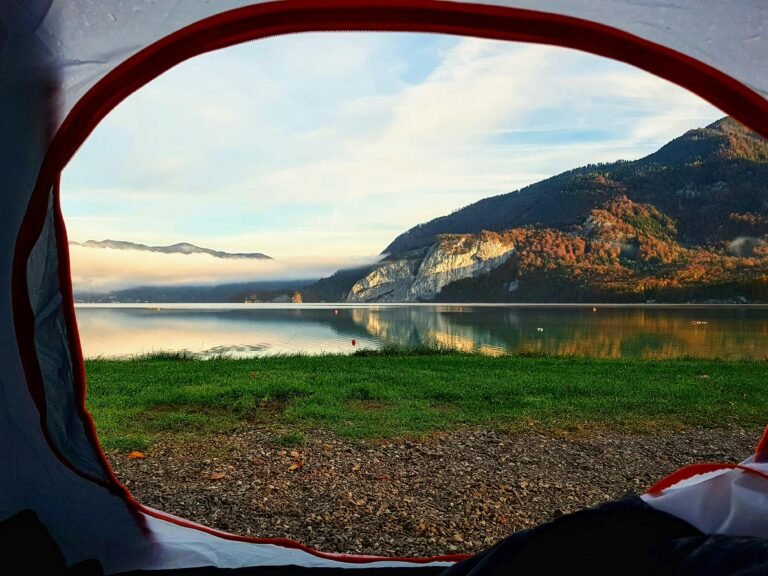 A peaceful view from inside a tent overlooking a calm lake surrounded by forested mountains. Soft sunlight reflects on the water, with mist hovering above the lake and autumn‑colored trees lining the mountains in the distance. Amazing Camping site