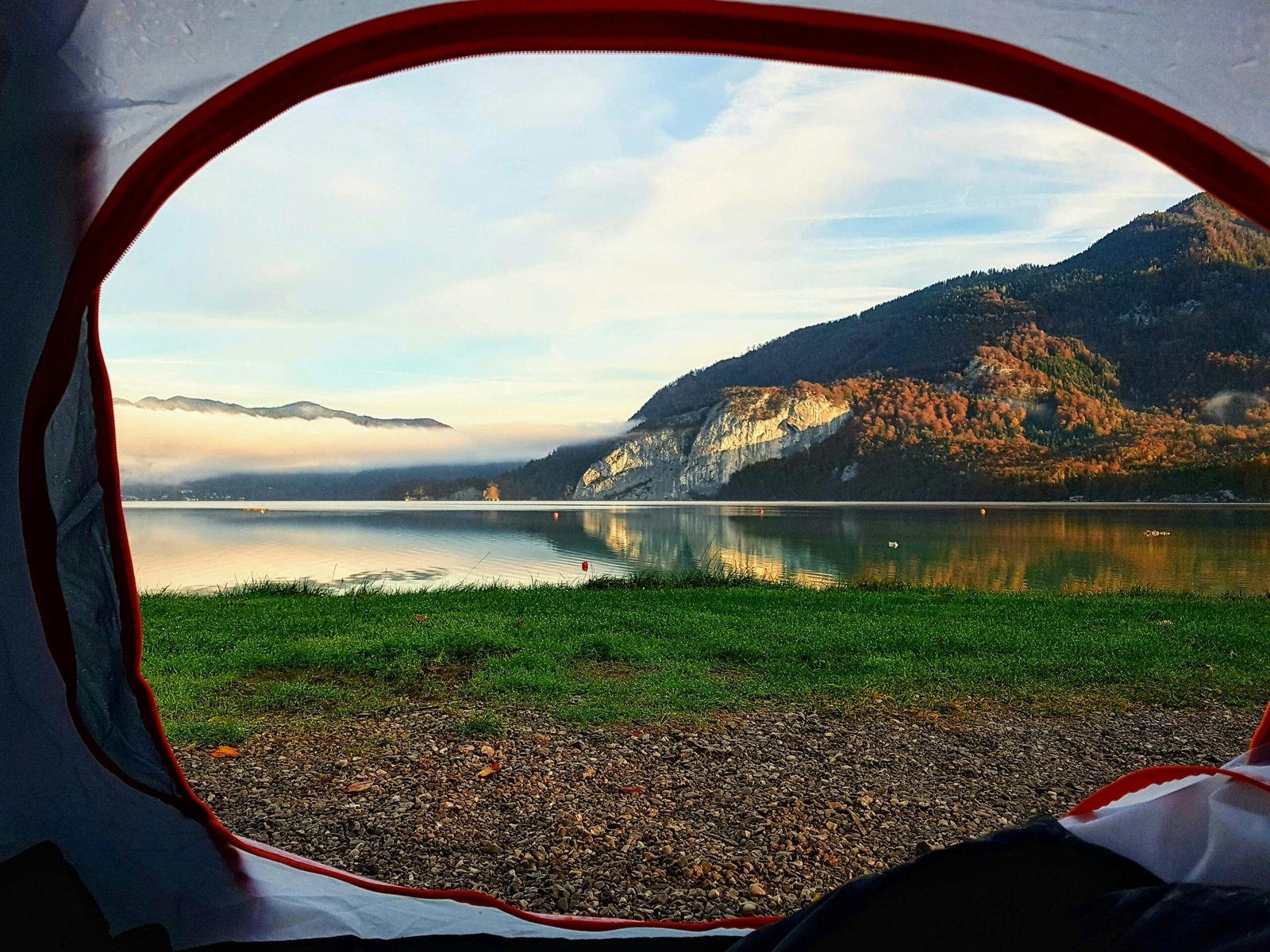 A peaceful view from inside a tent overlooking a calm lake surrounded by forested mountains. Soft sunlight reflects on the water, with mist hovering above the lake and autumn‑colored trees lining the mountains in the distance. Amazing Camping site