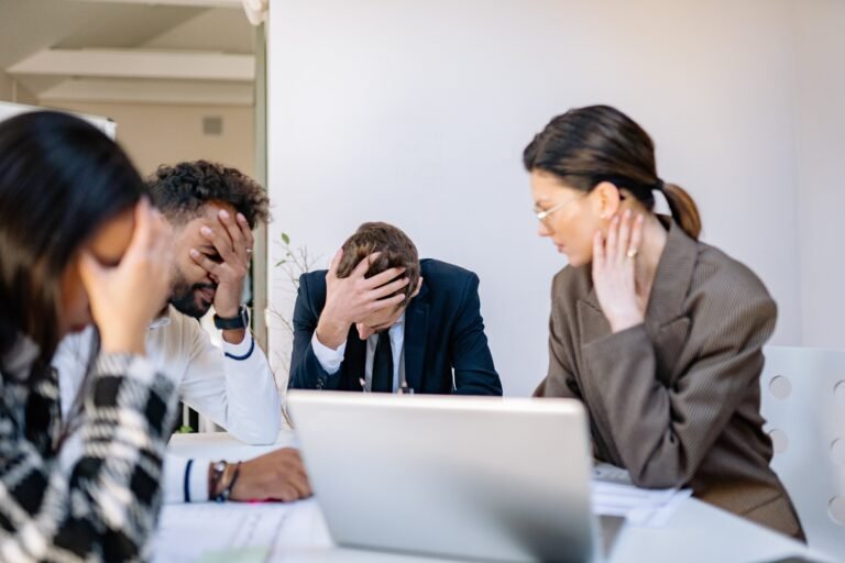 Four coworkers sit around a meeting table looking stressed and frustrated, with three holding their heads in their hands while documents and a laptop lie on the table during a tense discussion.
