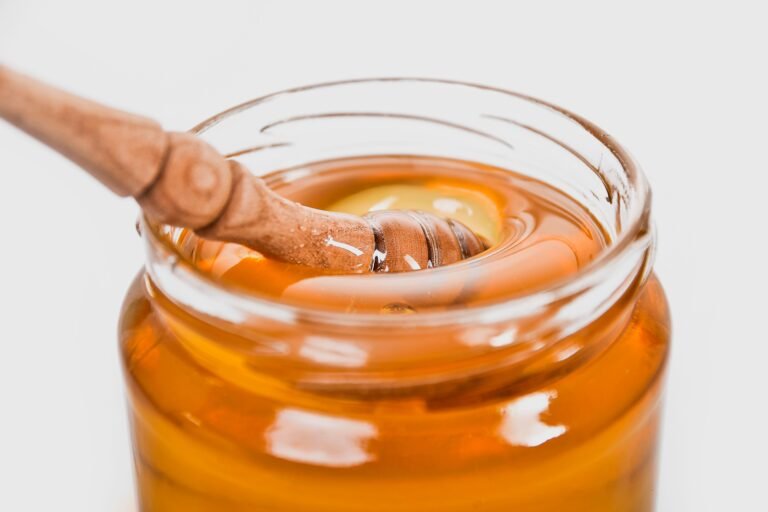 A glass jar filled with golden honey and a wooden honey dipper coated in thick, glossy honey, capturing its rich texture and natural shine.