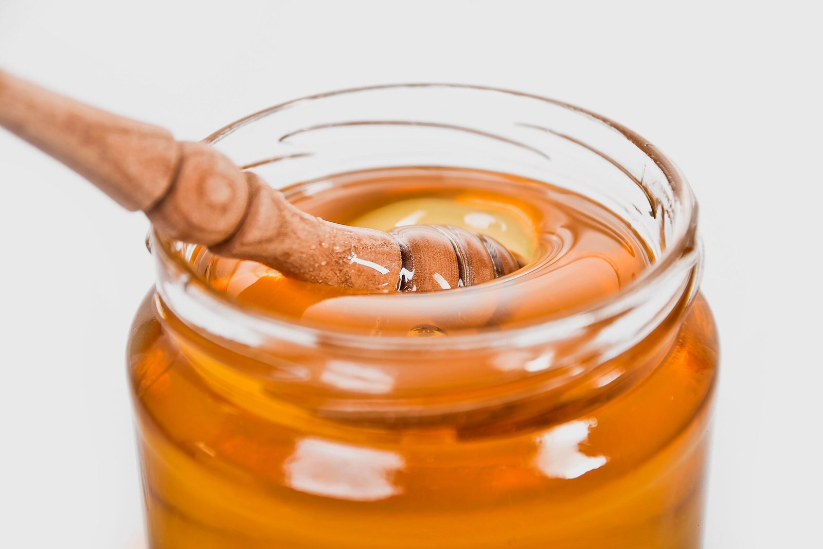 A glass jar filled with golden honey and a wooden honey dipper coated in thick, glossy honey, capturing its rich texture and natural shine.