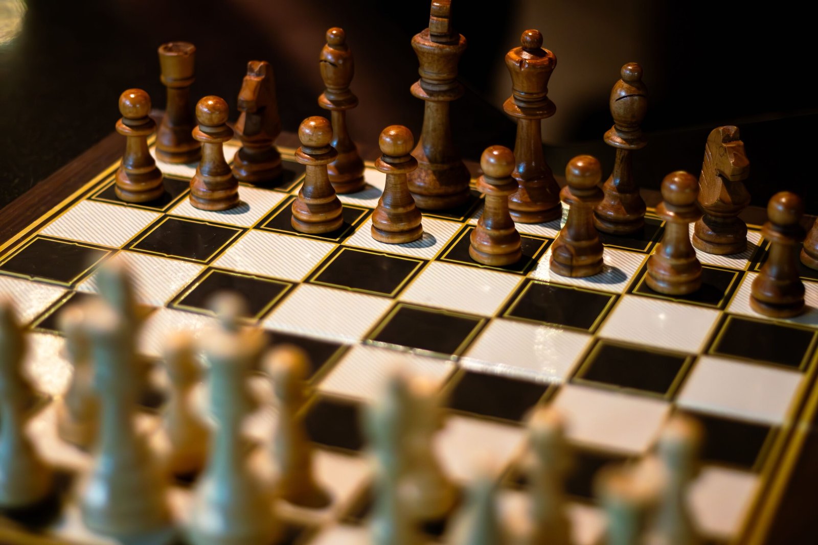 A close‑up view of a wooden chessboard with polished chess pieces arranged for the start of a game, with the dark pieces in focus and soft lighting highlighting the board’s texture.