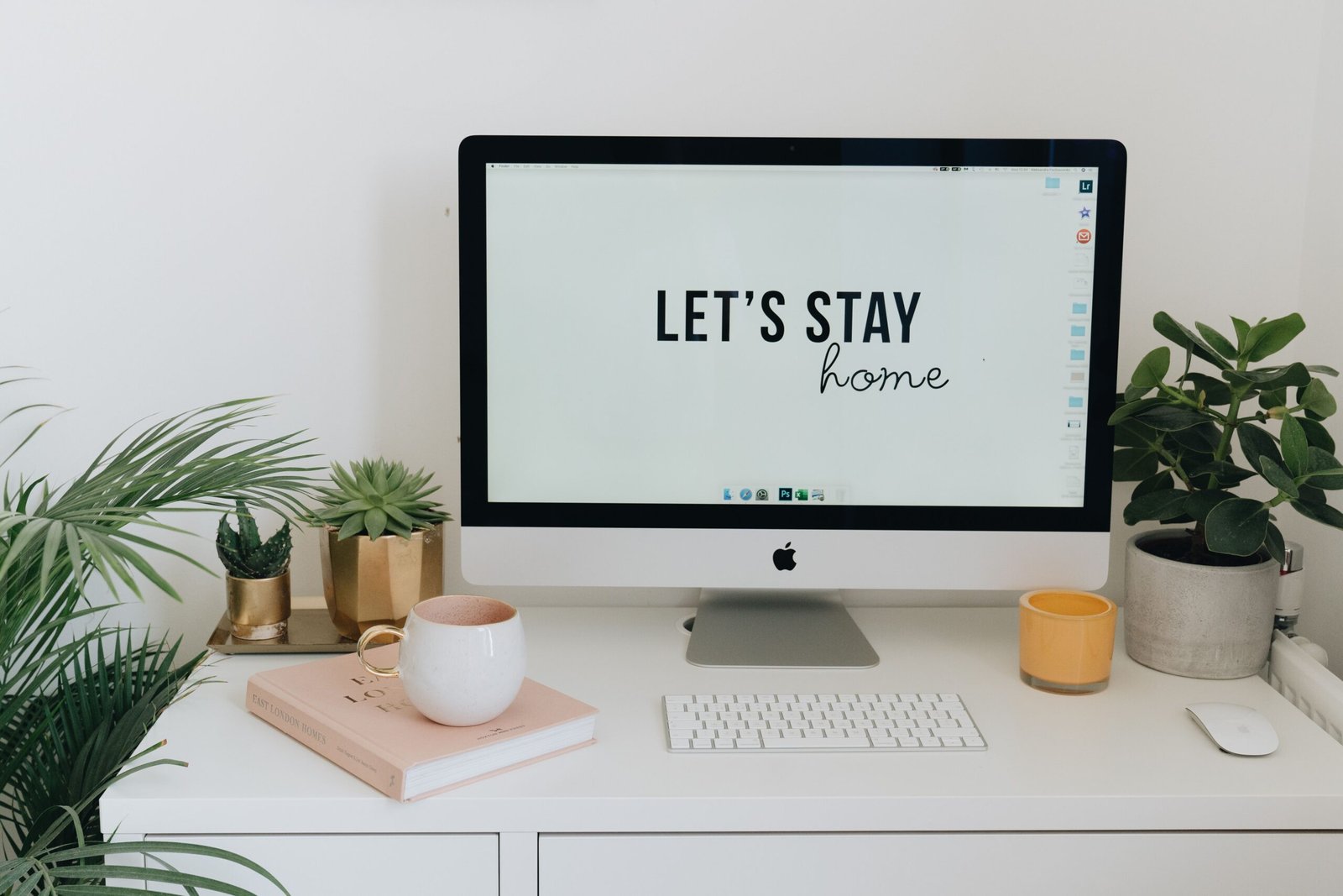 Cozy home office setup with an iMac displaying the words Lets Stay Home, surrounded by small potted plants, a pink book with a mug on top, a white keyboard and mouse, and a yellow candle on a bright white desk.