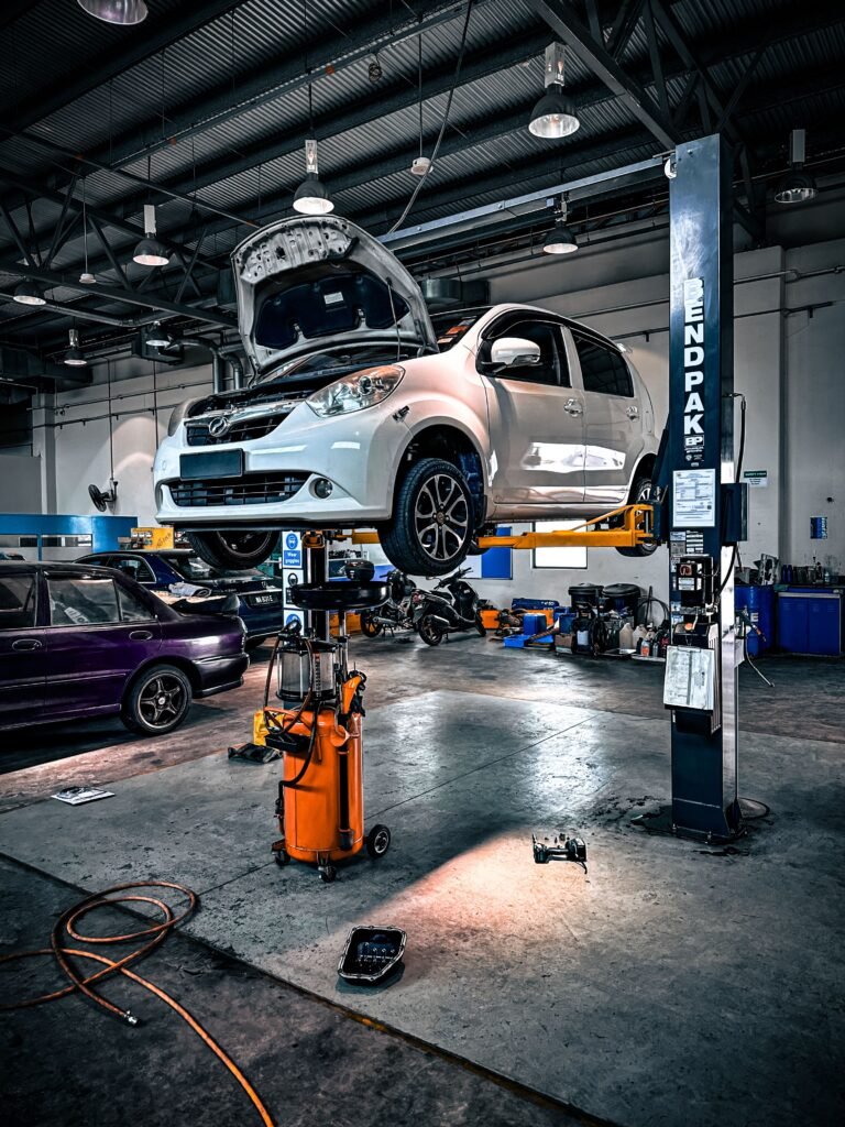 A white compact car lifted on a hydraulic service lift inside a professional auto repair shop, with the hood open and maintenance tools such as an oil drain tank and pans positioned underneath.