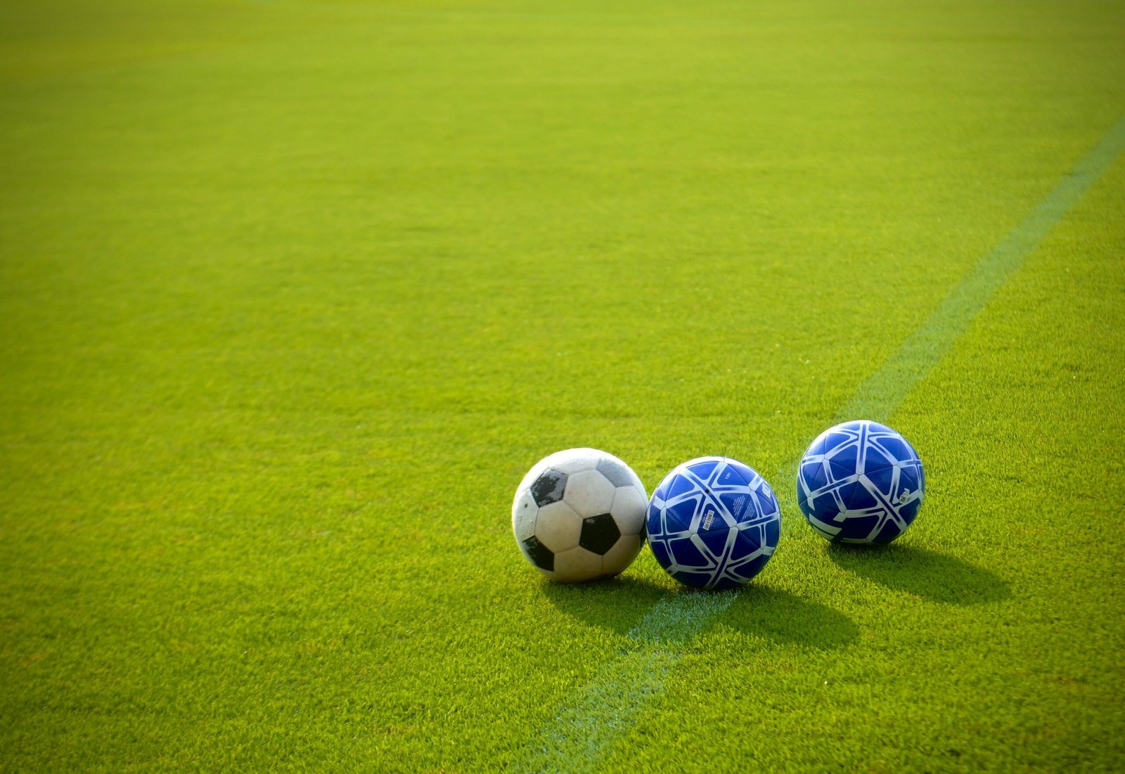 Three soccer balls resting on a green grass field, including one classic black‑and‑white ball and two modern blue‑and‑white designs, with a faint field line visible in the background.