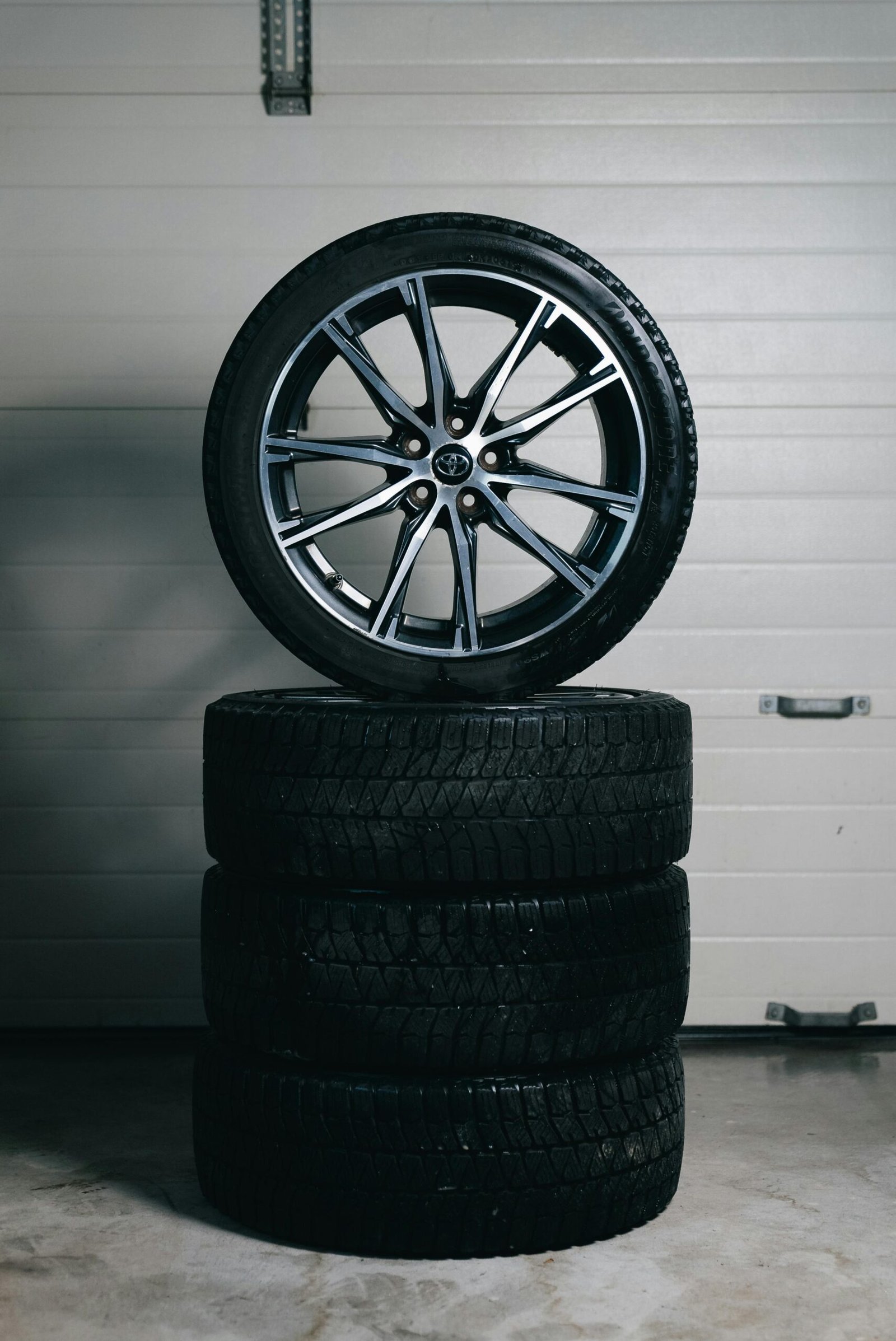 A stack of four car tyres with a multi‑spoke Toyota alloy wheel placed on top, positioned on a concrete garage floor in front of a closed garage door.