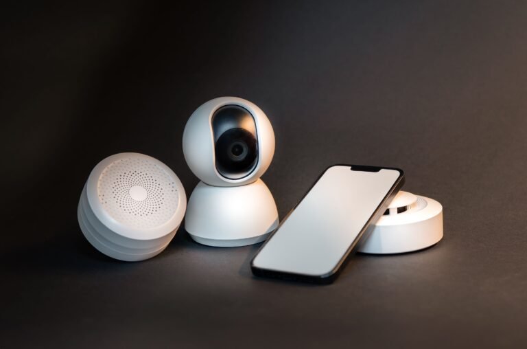 Smart home devices arranged on a dark surface, including a smart security camera, a round smart speaker, a smart sensor, and a smartphone used to control the devices