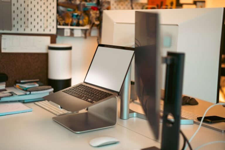 A modern home office setup with a laptop on a metallic stand in front of two large monitors on a clean desk with notebooks, papers, and a corkboard in the background.
