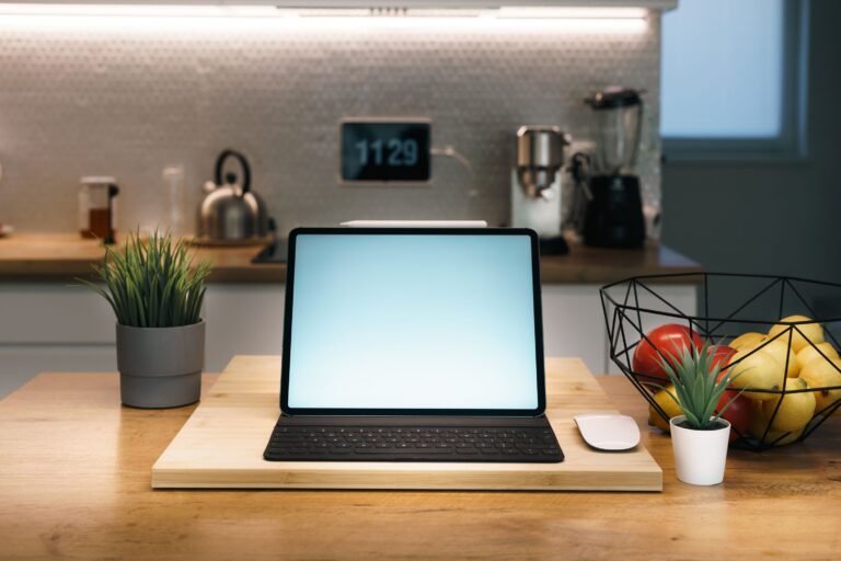 Modern kitchen countertop with a tablet on a wooden stand, wireless mouse, small potted plants, and a fruit basket with lemons and tomatoes, with kitchen appliances in the background.