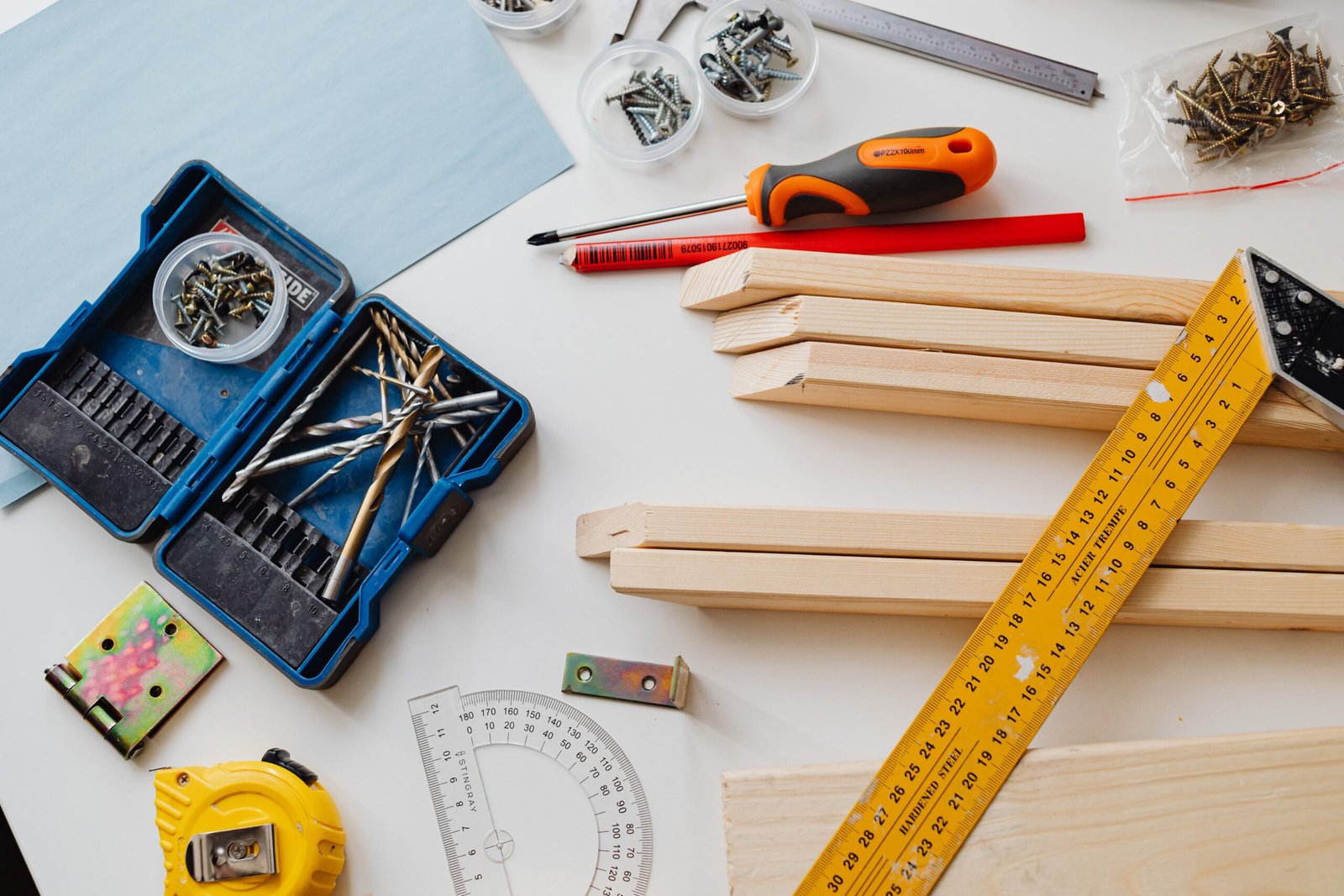 A neatly arranged DIY workspace with woodworking tools, including a drill bit set, screws, a screwdriver, measuring tape, rulers, a pencil, metal brackets, and pieces of wood on a tabletop.