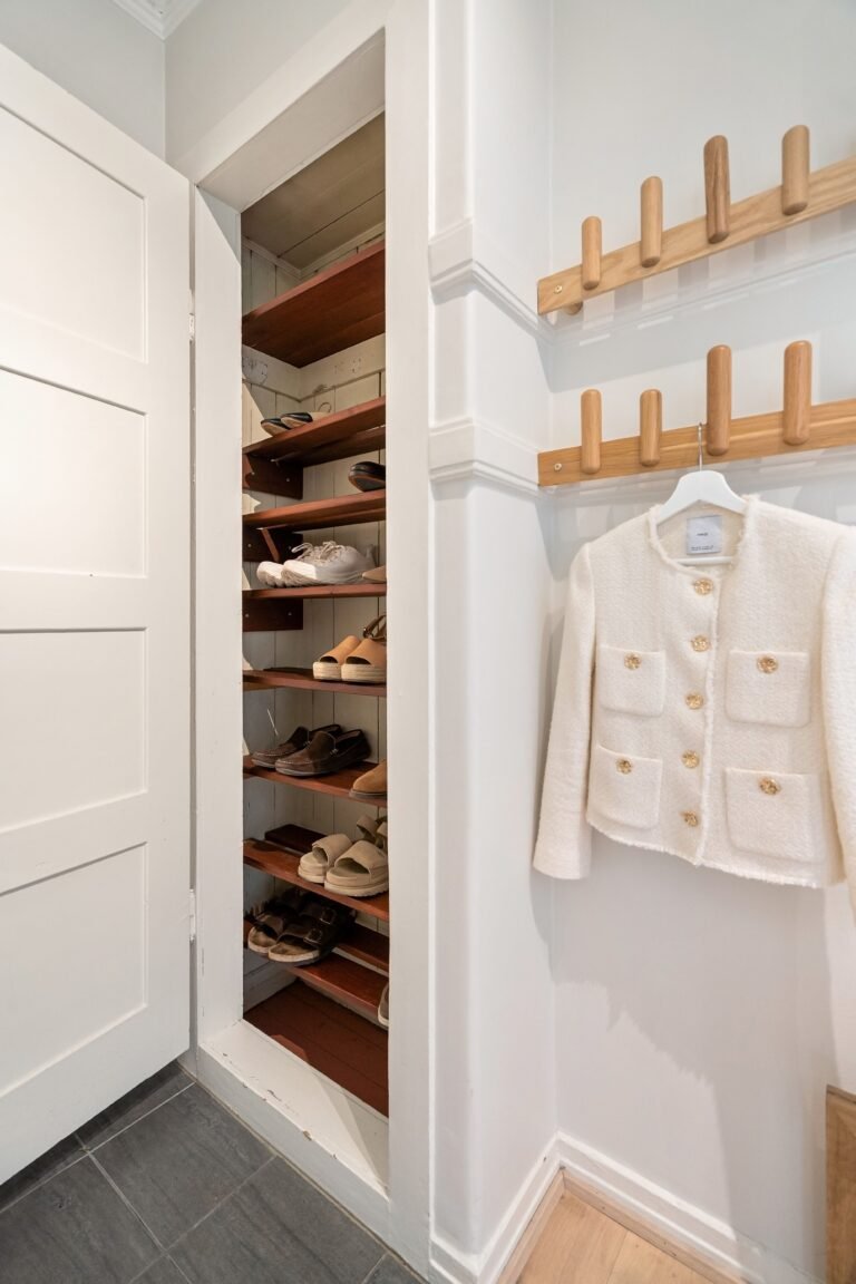 Small organized closet with wooden shelves holding neatly arranged shoes, next to wall mounted coat racks with a white textured jacket hanging above light wood flooring.