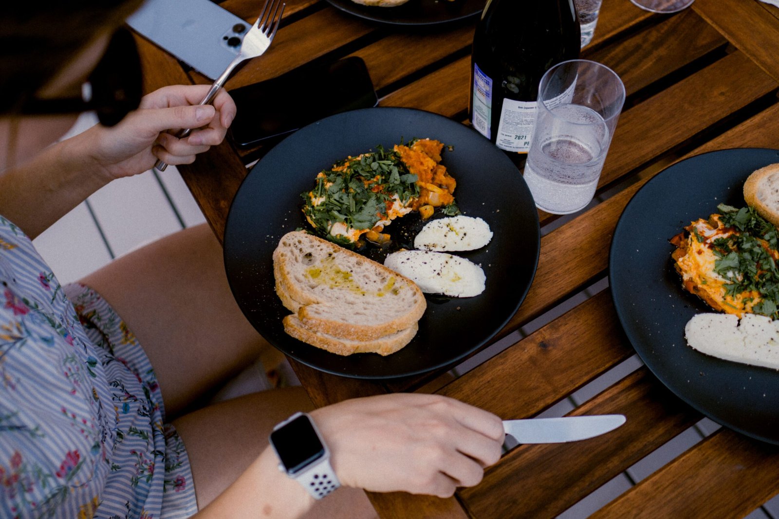 Person eating a meal at an outdoor wooden table with bread drizzled with olive oil, white cheese sprinkled with pepper, and a vegetable egg dish, with a glass of water and a bottle of sparkling drink nearby.