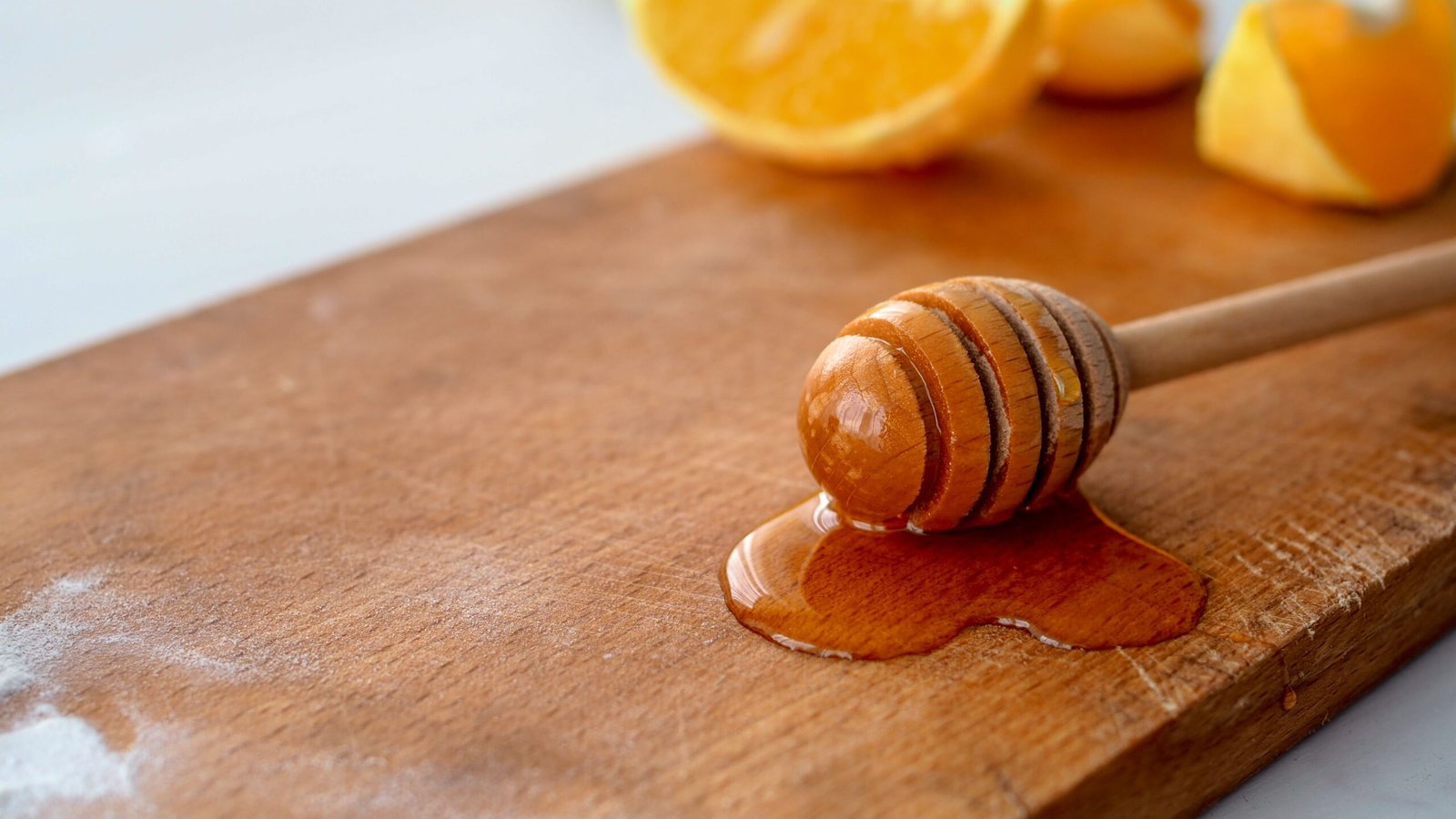 A wooden honey dipper resting on a wooden surface with golden honey dripping from it, with sliced citrus fruit softly blurred in the background for a fresh, natural look.