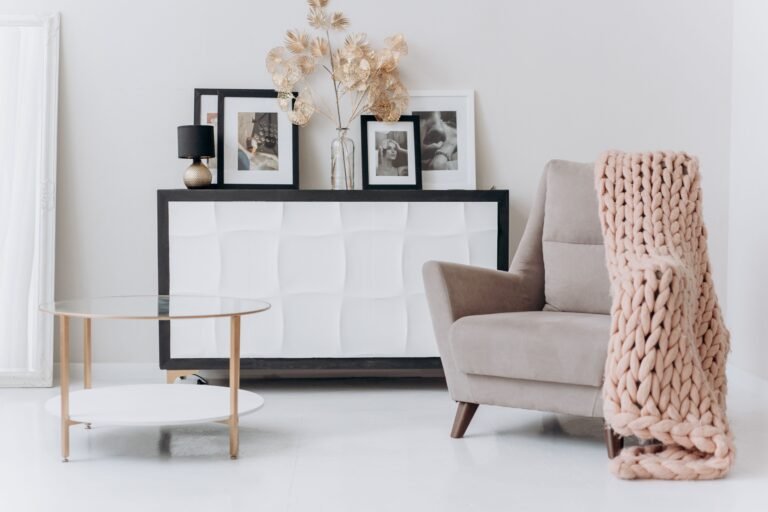 A modern, minimalist living room corner featuring a beige armchair with wooden legs, draped with a blush pink chunky knit blanket. A round glass coffee table with gold legs sits beside it, and a white cabinet with a textured front holds framed art, a small lamp, and a vase with gold decorative leaves. A large white-framed mirror leans against the wall, creating a bright, cozy, elegant atmosphere.