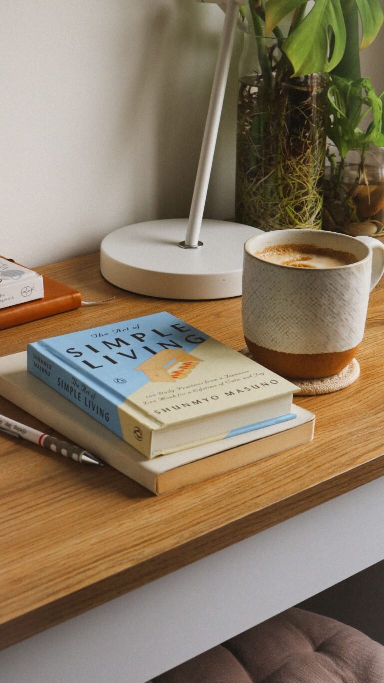 A cozy minimalist desk setup with a coffee mug, stacked books, a white lamp, a pen, and a glass vase with green plants on a wooden surface.