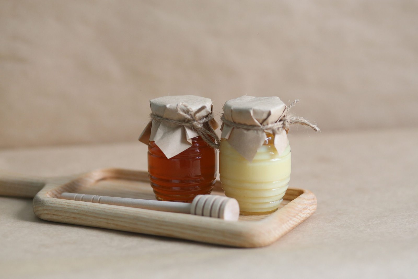 Two small glass jars filled with different types of honey, one dark and one light, covered with brown paper and twine, displayed on a wooden tray with a honey dipper for a natural, rustic look.