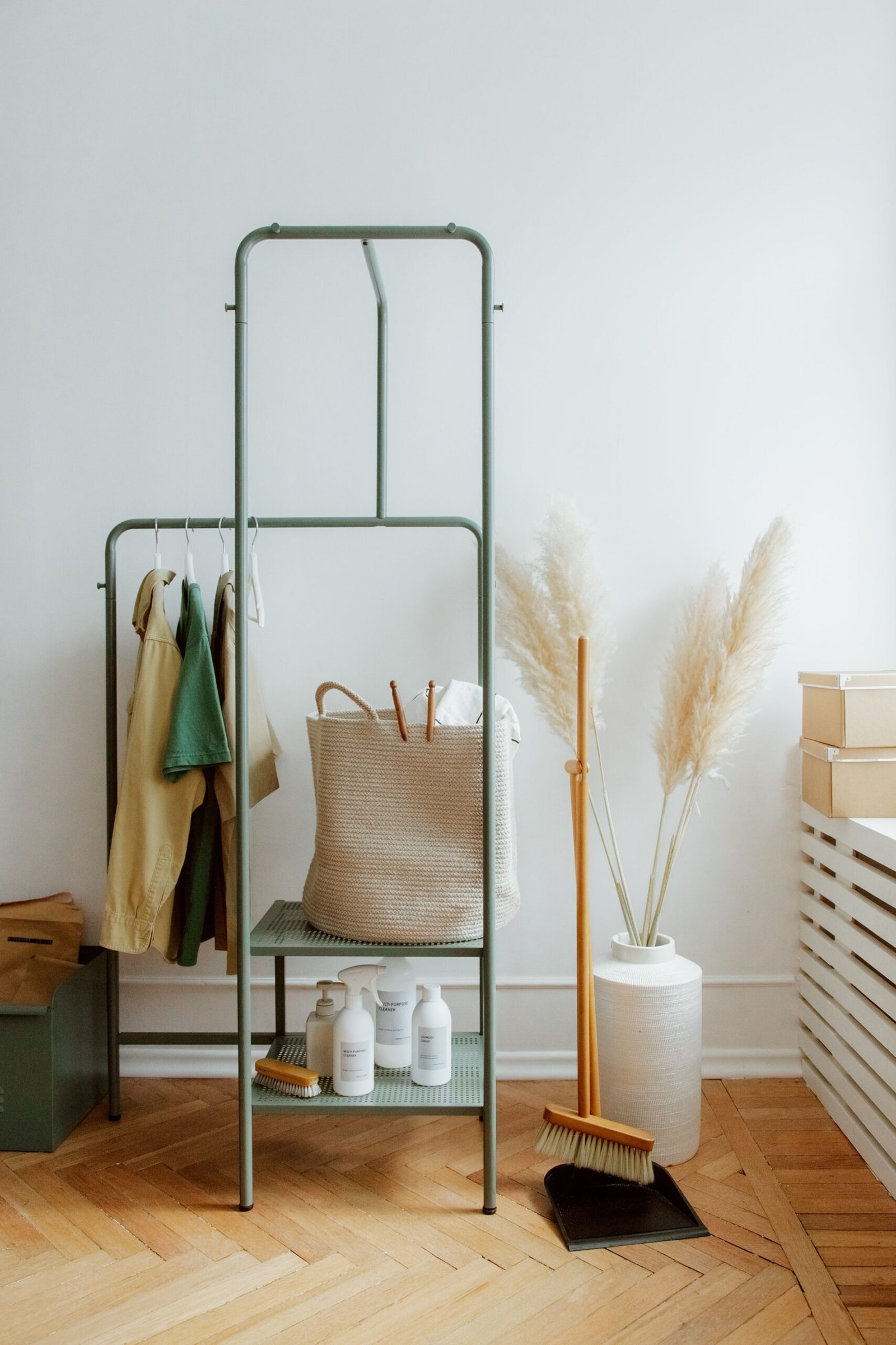 Minimalist home organization setup with a green metal rack holding neatly hung clothes, a woven basket, white storage bottles, a vase with pampas grass, and stacked boxes on a bench in a bright and tidy room.