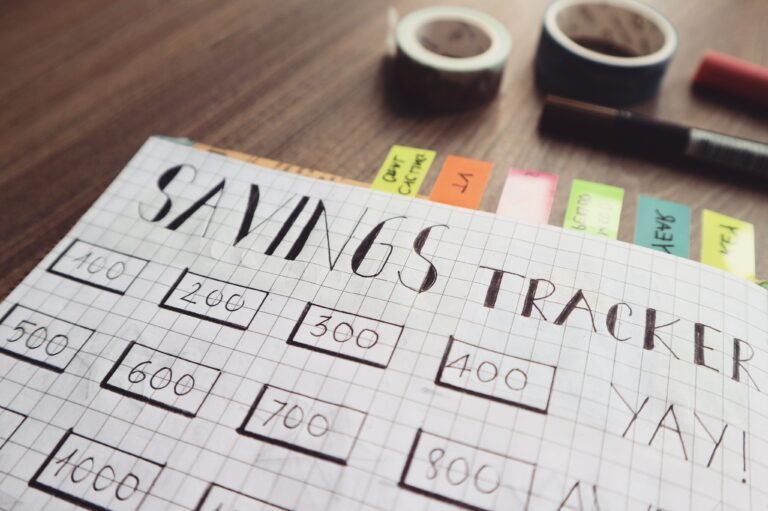 Hand drawn savings tracker on graph paper with numbered boxes from one hundred to one thousand, surrounded by colorful sticky notes, decorative tape, and a black marker on a wooden table.