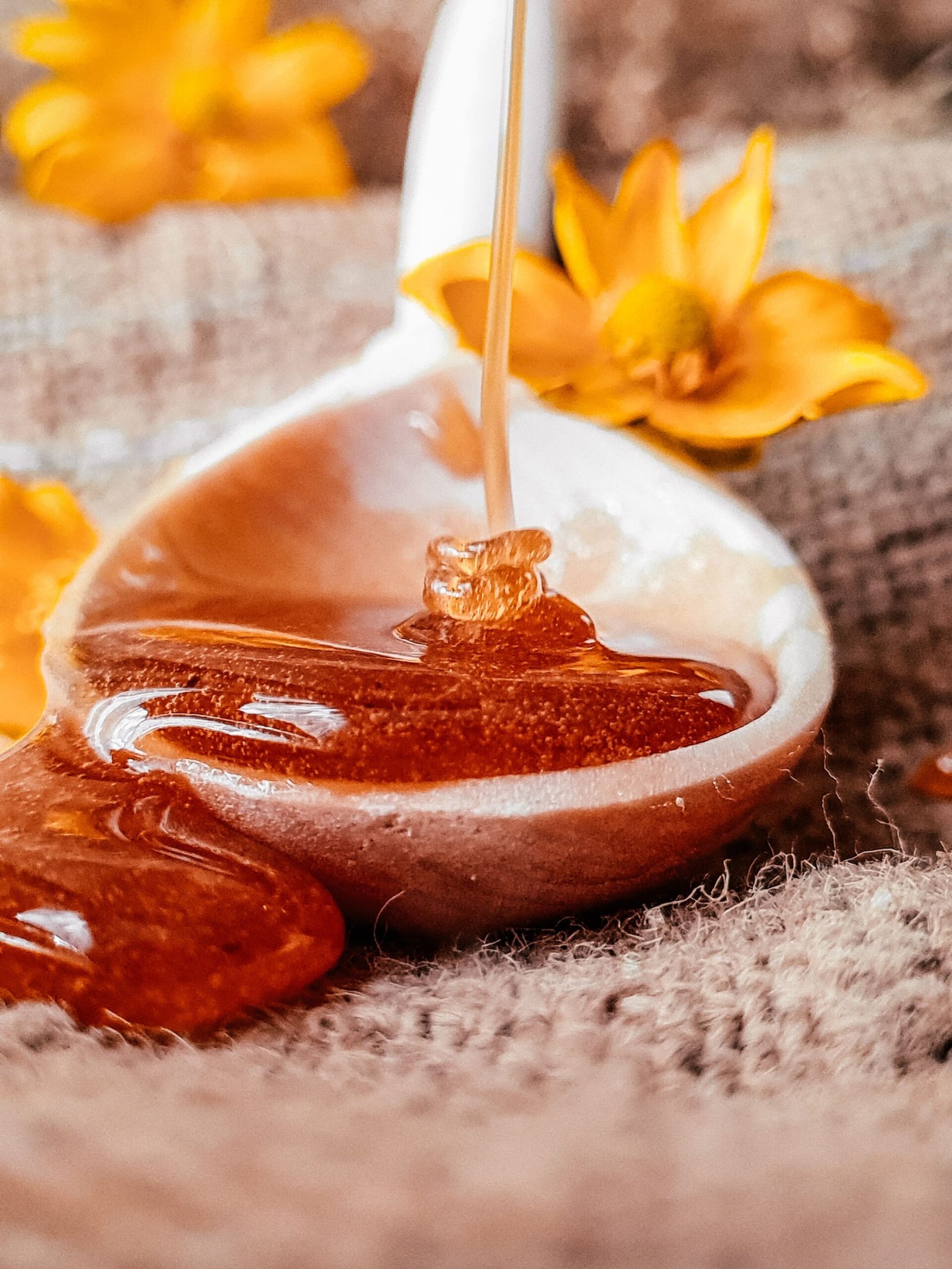 A wooden spoon filled with golden honey as it pours smoothly onto a burlap surface, surrounded by bright yellow flowers for a warm and natural look.