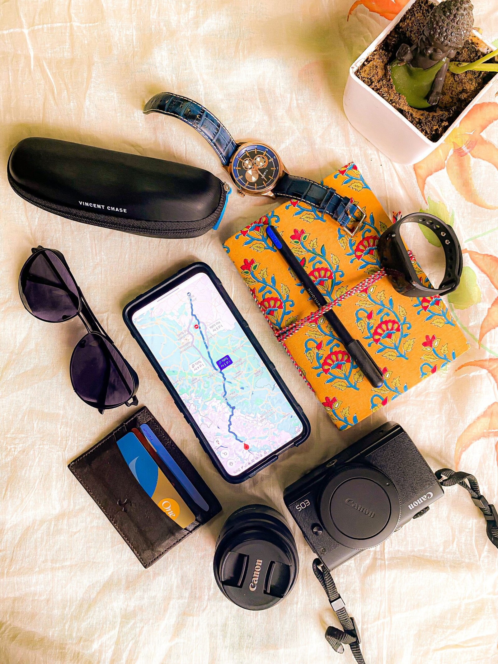 A neatly arranged flat lay of travel essentials including sunglasses, a wristwatch, a smartphone showing a map, a floral notebook with a pen, a fitness band, a wallet with cards, a Canon camera, and a small Buddha plant on a light fabric background.