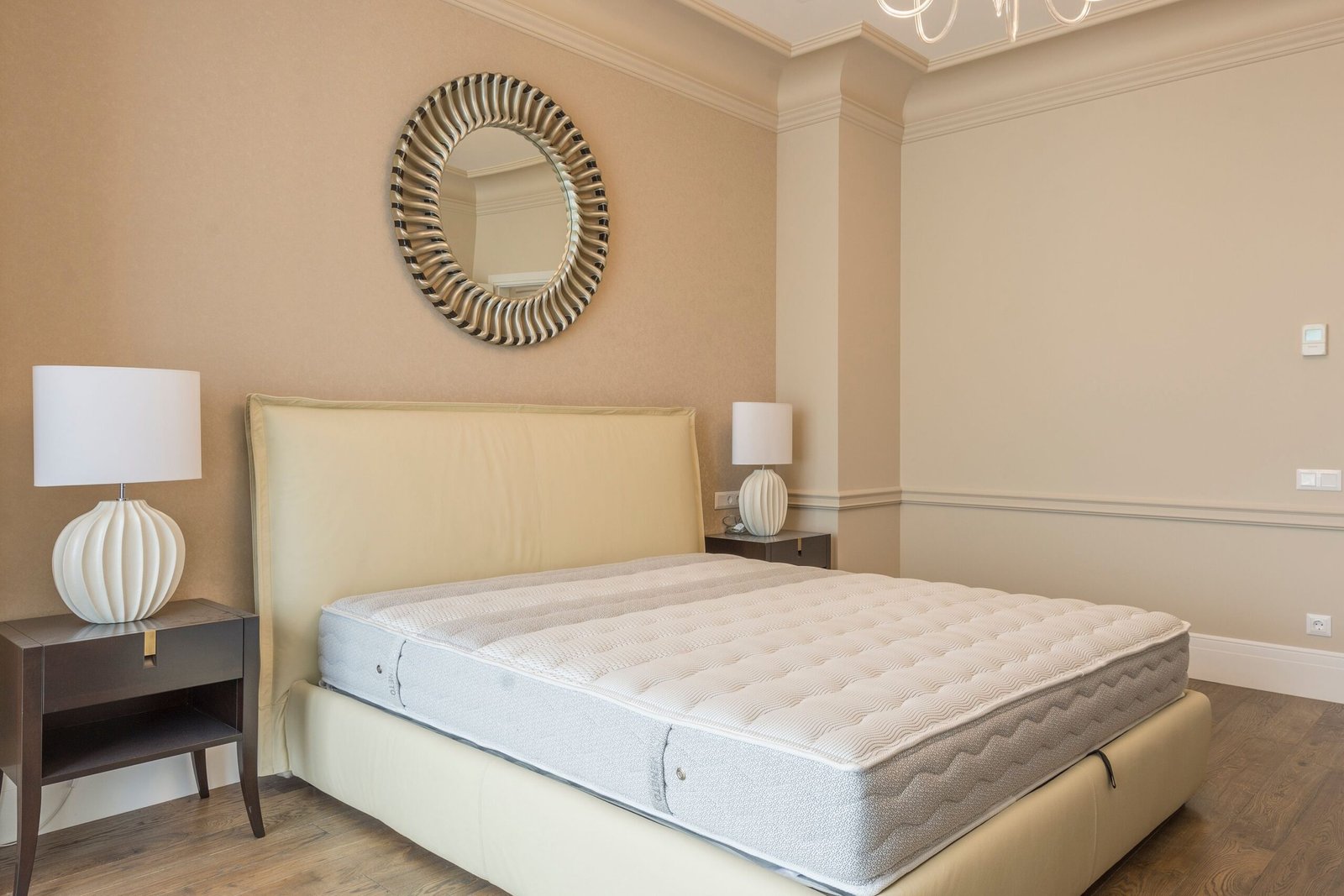 A modern bedroom featuring a beige upholstered headboard, a bare mattress, and matching ceramic lamps on dark wooden nightstands. A round metallic mirror hangs above the bed, creating a calm and elegant atmosphere.