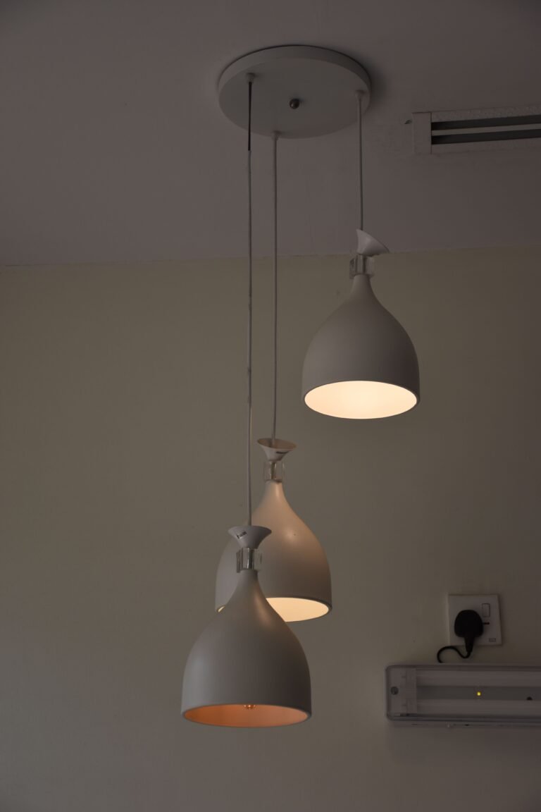A set of three modern pendant lights hangs from the ceiling at staggered heights. Each light has a smooth matte white finish, with two lamps glowing warmly while one remains off. A nearby wall shows an electrical outlet with a connected plug and a small mounted fixture, creating a simple and minimalist interior lighting scene.