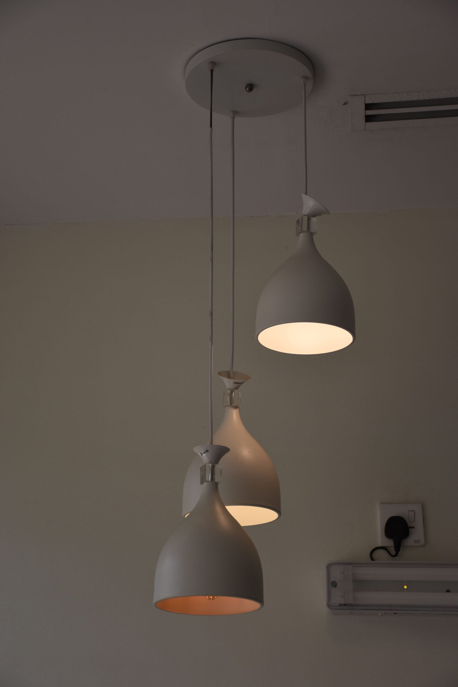 A set of three modern pendant lights hangs from the ceiling at staggered heights. Each light has a smooth matte white finish, with two lamps glowing warmly while one remains off. A nearby wall shows an electrical outlet with a connected plug and a small mounted fixture, creating a simple and minimalist interior lighting scene.