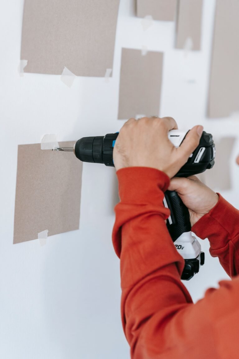 Person using a cordless drill to drive a screw into a white wall, with paper templates taped up to plan where frames will hang