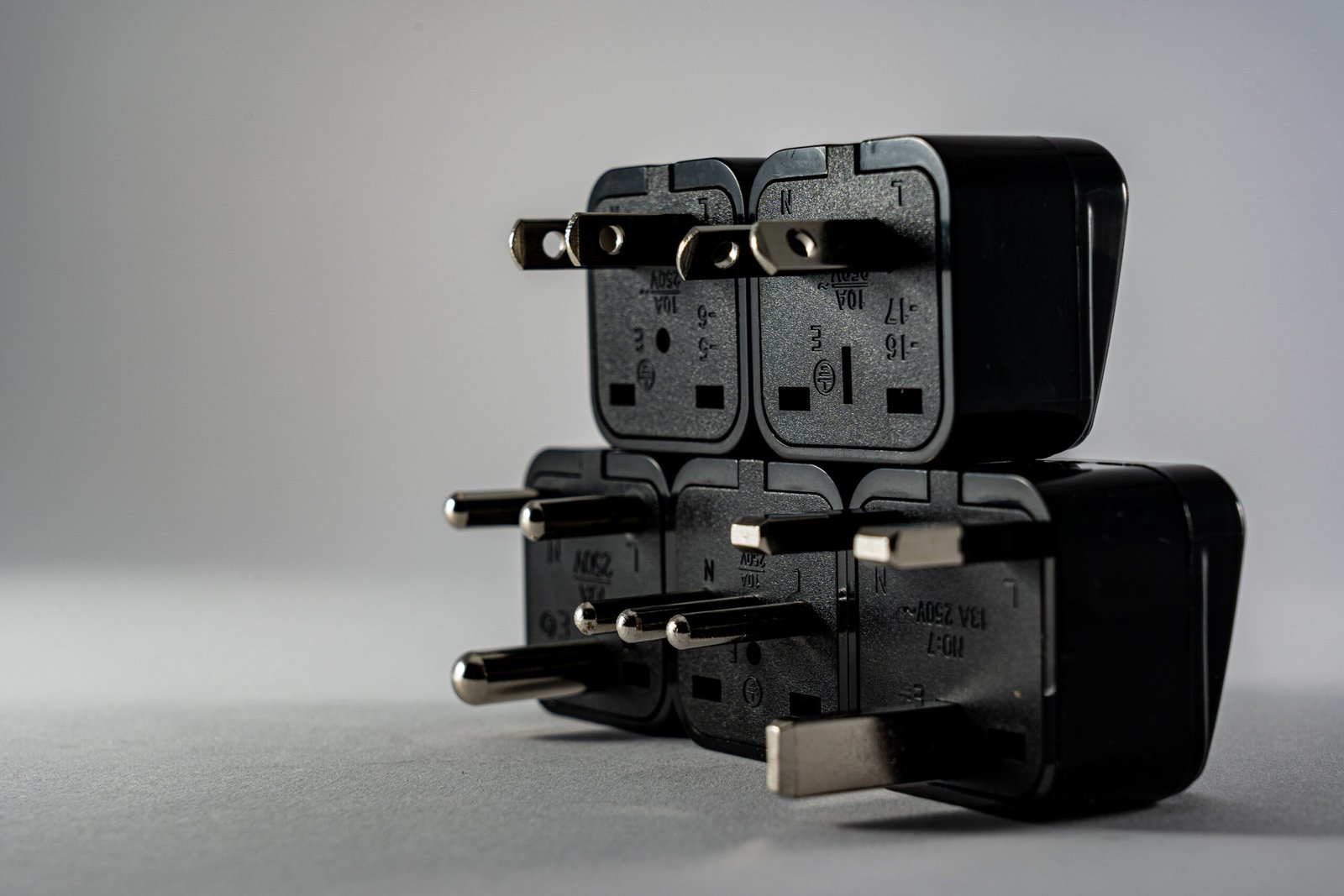 Stack of black travel plug adapters with different prong styles, including flat, round and rectangular pins, on a grey surface