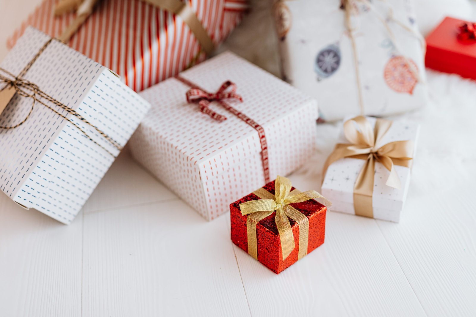 Christmas gifts wrapped in red striped and white patterned paper with gold and red ribbons, with a small sparkly red box at the front, on a white wooden floor