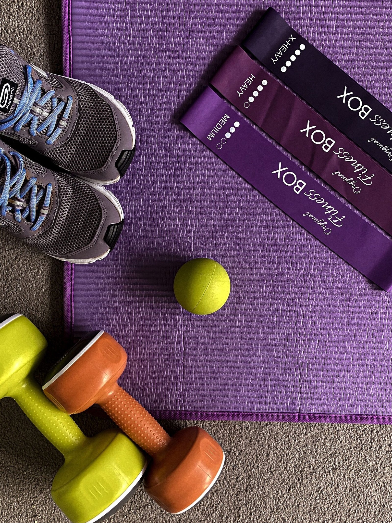 Three purple resistance loop bands labelled medium, heavy and extra heavy on a purple exercise mat, with sneakers, a massage ball and two small dumbbells