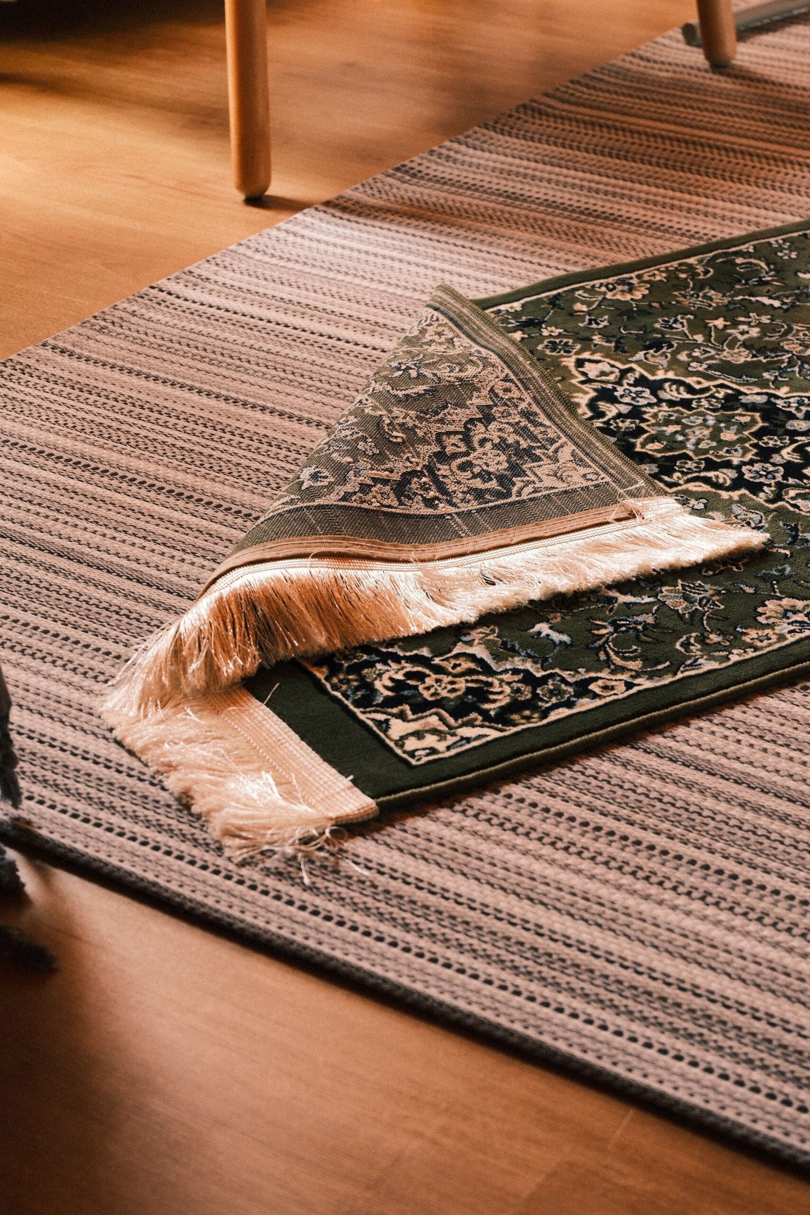 Small green patterned rug with a folded fringed corner layered on top of a larger striped flat woven rug on a wooden floor