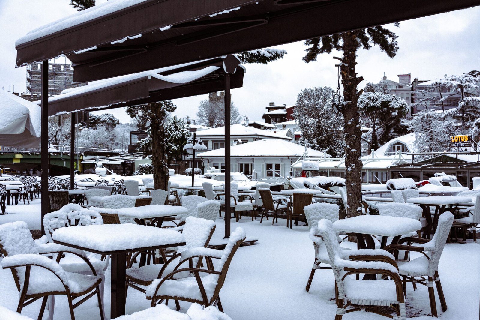 Outdoor patio tables and wicker chairs covered in thick snow under a snow laden canopy, with snowy trees and buildings behind