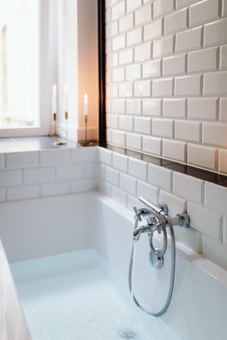 White bathtub filling with water from a chrome faucet, with white subway tile above the tub edge and candles glowing on a windowsill
